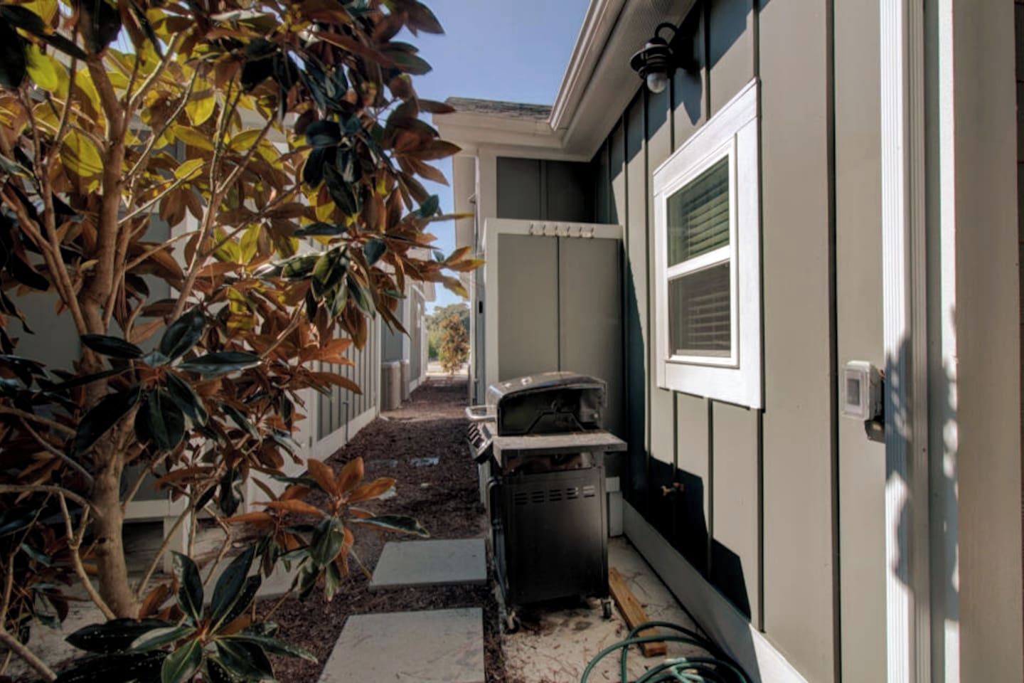 A narrow pathway leading to a house with a trash bin and a hose on the ground.