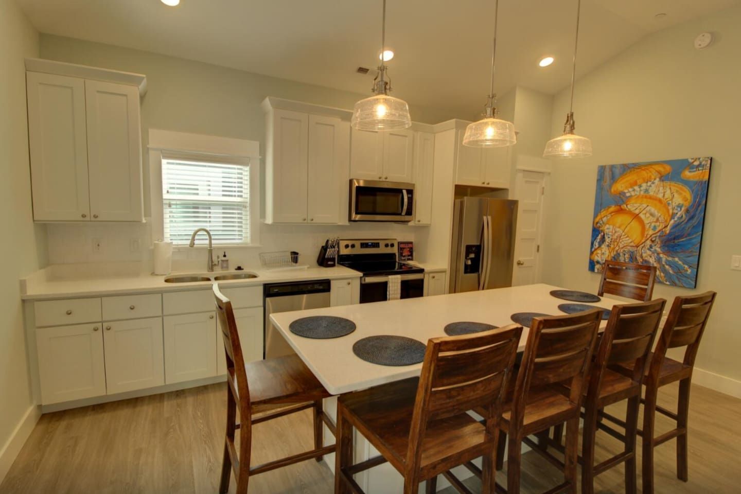 A modern kitchen with white cabinets, a central island with chairs, a microwave, a refrigerator, and pendant lighting.
