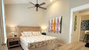 A bedroom with a bed, a nightstand, a table, a ceiling fan, and artwork on the walls. - Dog Friendly Luxury Condo, Walk to the Beach (Corolla)
