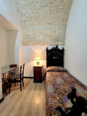 Un dormitorio con pared de piedra, una cama con colcha estampada, una mesita de noche con lámpara, una silla y una pequeña estatua sobre la mesita.