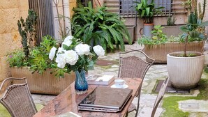 Pátio externo com mesa de madeira e cadeiras de metal, cercado por plantas em vasos e piso de pedra.