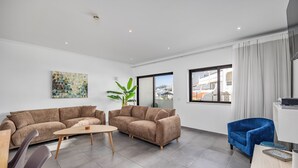 Una sala moderna con dos sofás, una mesa de centro y un sillón azul. Hay un ventanal con vistas a edificios y una planta en la esquina.