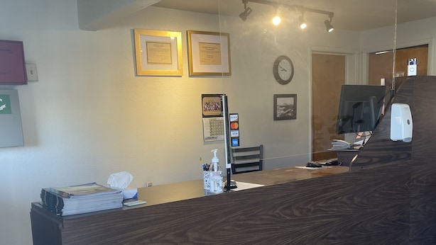 A reception desk with a dark wood finish, a computer monitor, and a payment terminal. There are framed certificates on the wall and a clock. - CRYSTAL MOTEL (Red Bluff)