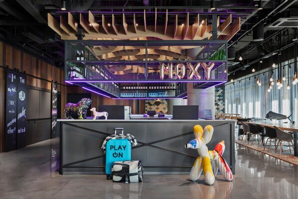 A modern reception area with a neon sign reading "MIX" and a counter with a digital display. There are luggage bags and a colorful sculpture on the floor.