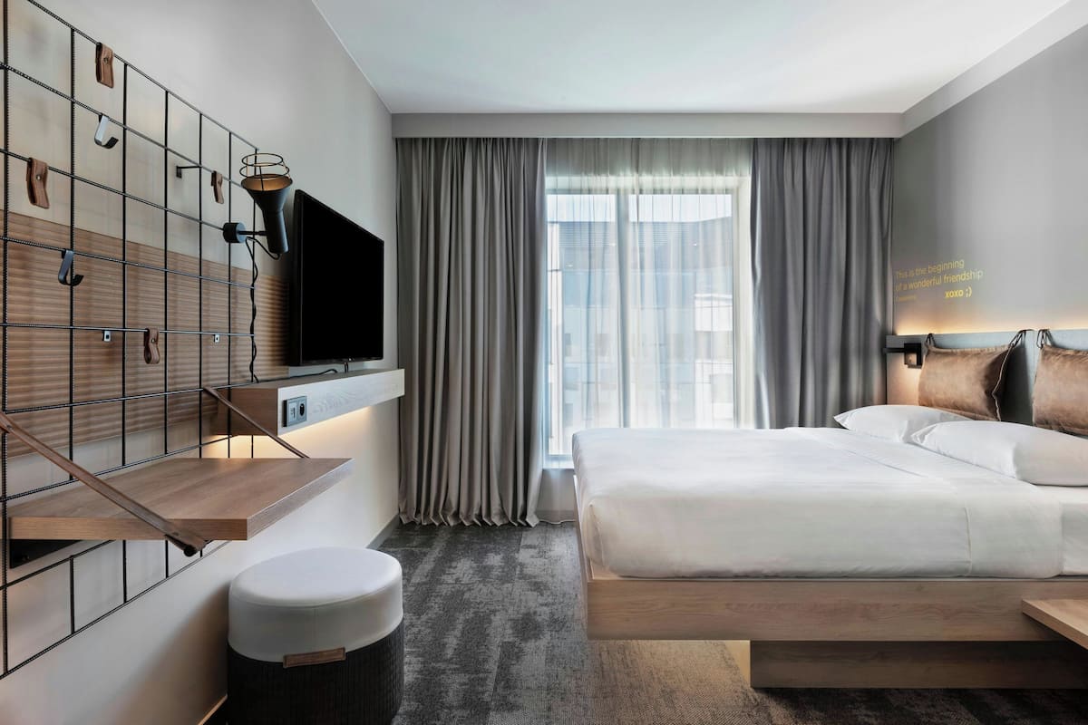 Une chambre d’hôtel moderne avec un grand lit, un téléviseur fixé au mur, une étagère en bois et une grande fenêtre avec des rideaux.
