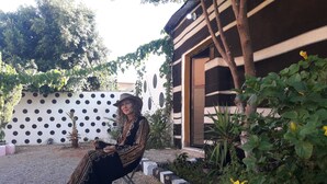 Una persona sentada en un taburete en un jardín con un muro blanco con puntos negros, un muro a rayas blancas y negras, y una flor amarilla.