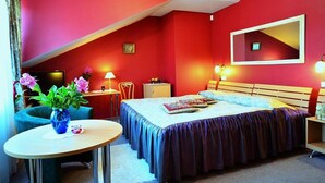 A bedroom with a red wall, a bed with a blue bedspread, a small table with a vase of flowers, and a chair. - Centro Kubas - Angel (Vilnius)