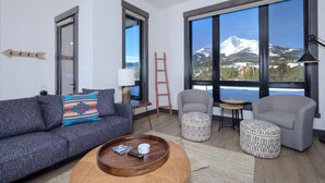 Een moderne woonkamer met een grijze bank, een salontafel en twee voetenbankjes. Grote ramen bieden uitzicht op een berg en een meer.