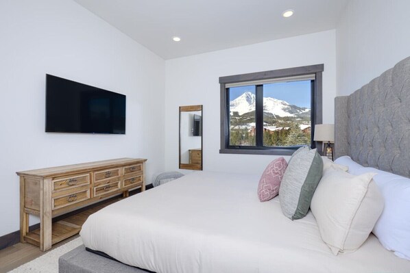 A bedroom with a large bed, a wooden dresser, a flat-screen TV, a window with a mountain view, and a mirror. - Magnificent Home, Views of Lone Peak, Hot Tub (Big Sky)