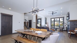 Een ruime woonkamer met een houten eettafel, banken en een moderne kroonluchter. Een trap leidt naar een hoger niveau, er is een open haard en een groot raam met uitzicht op de omgeving.