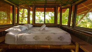 A bed with a floral bedspread, pillows, and a rolled towel in a room with bamboo walls and large windows. - Bamboo Beach House Steps away from secluded beach (Provincia de Puntarenas)