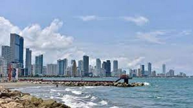 A coastal cityscape with modern high-rise buildings, a rocky shoreline, and calm sea.