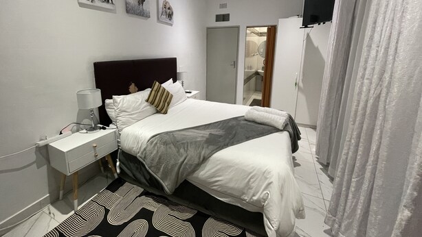 A modern bedroom with a large bed, a bedside table, a mirror, and a shower area visible through an open door. - SEMENI ASANTE GUESTHOUSE (Johannesburg South)