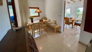 Een ruime woonkamer met een houten eettafel, een bank en een lange bank. Er is een raam met gordijnen, een hanglamp en een plafondventilator.