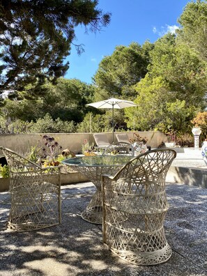 An outdoor seating area with wicker chairs and a glass table, surrounded by trees and a concrete wall. - Vacation home with ocean views and a large lot right on Playa Migjorn (Platja de Migjorn)