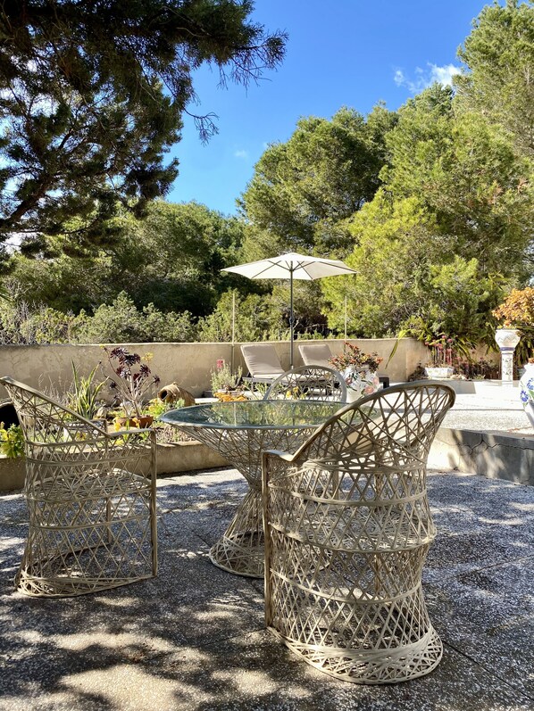 An outdoor seating area with wicker chairs and a glass table, surrounded by trees and a concrete wall. - Vacation home with ocean views and a large lot right on Playa Migjorn (Platja de Migjorn)