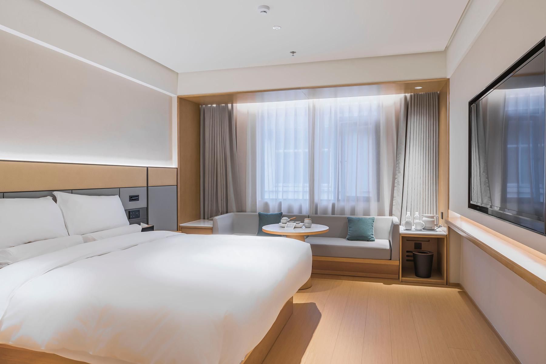 Una habitación de hotel con una cama grande, una zona de estar con una mesa y un ventanal con cortinas.
