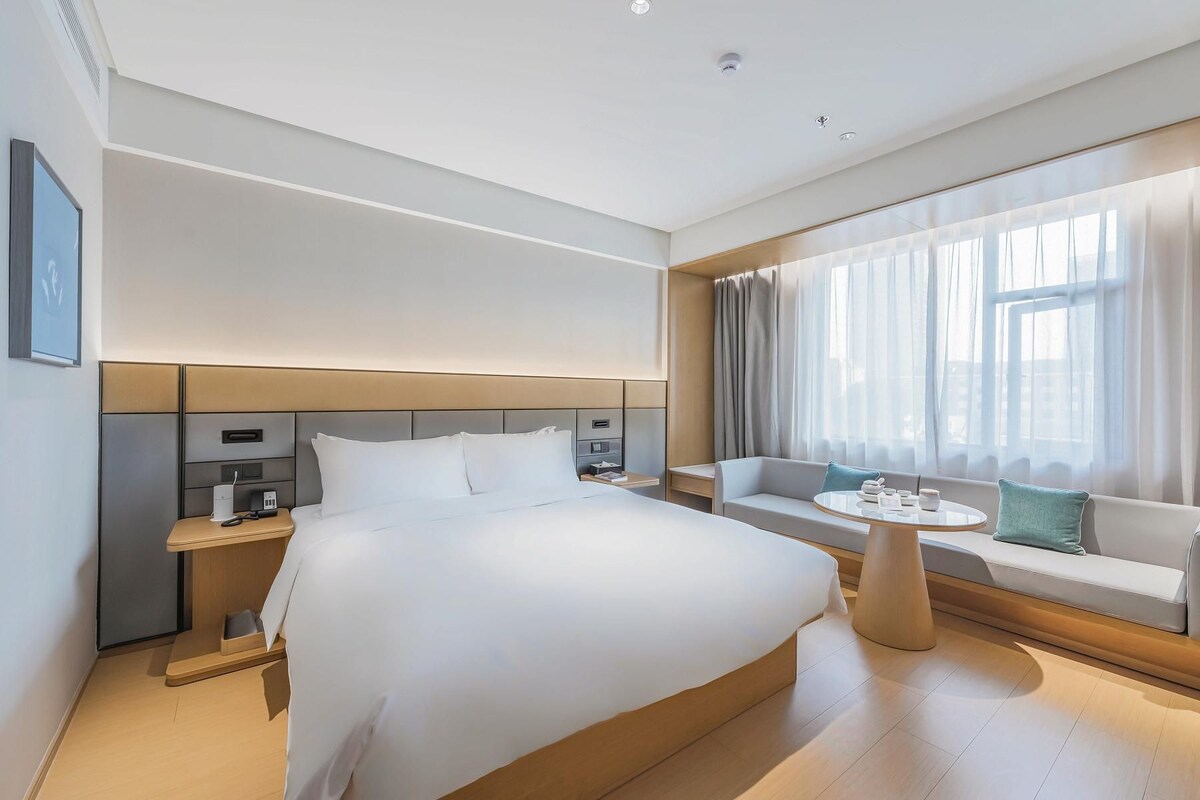 A modern hotel room with a large bed, bedside tables, a seating area with a table, and a large window with curtains.