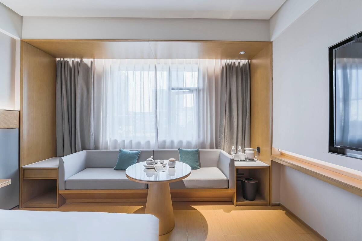 A hotel room with a sofa, a small round table, a trash bin, and a window with curtains.