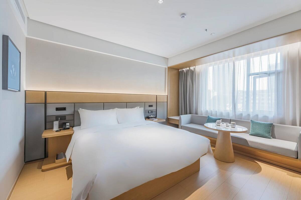 A modern hotel room with a large bed, bedside tables, a seating area with a table, and a large window with curtains.