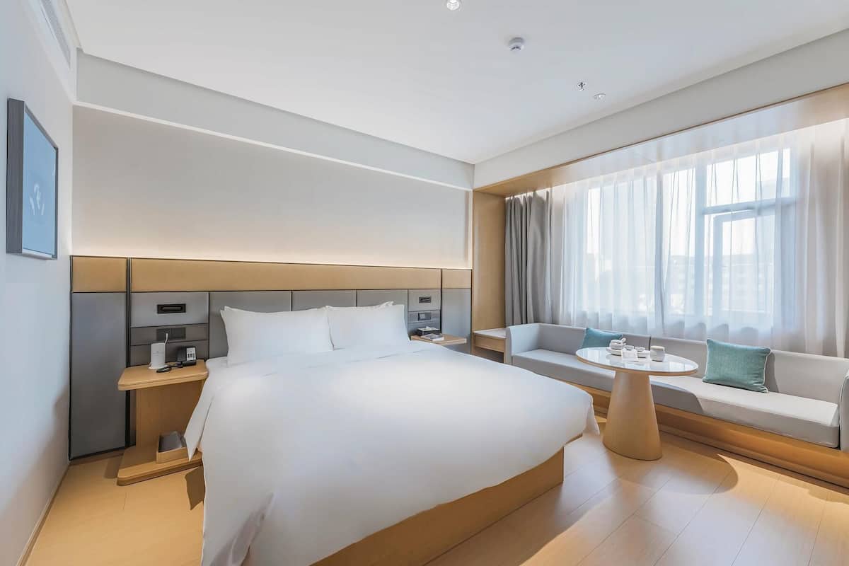 A modern hotel room with a large bed, bedside tables, a seating area with a table, and a large window with curtains.