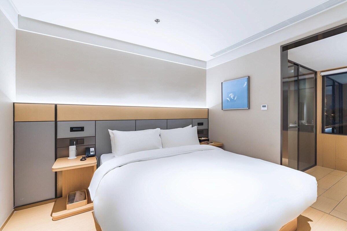 A modern hotel room with a large bed, bedside tables, a nightstand, a wall-mounted control panel, and a sliding glass door leading to a balcony.