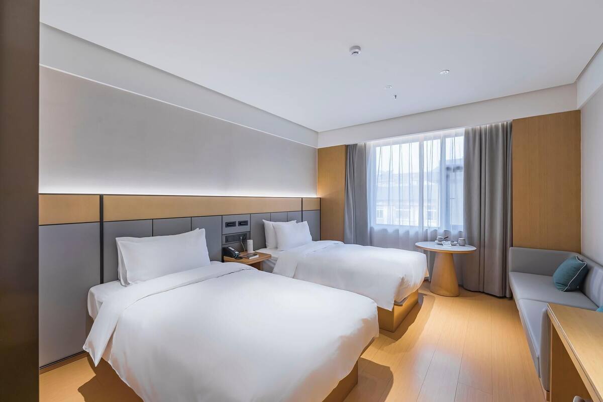 A hotel room with two beds, a small table, a sofa, and a large window with curtains.