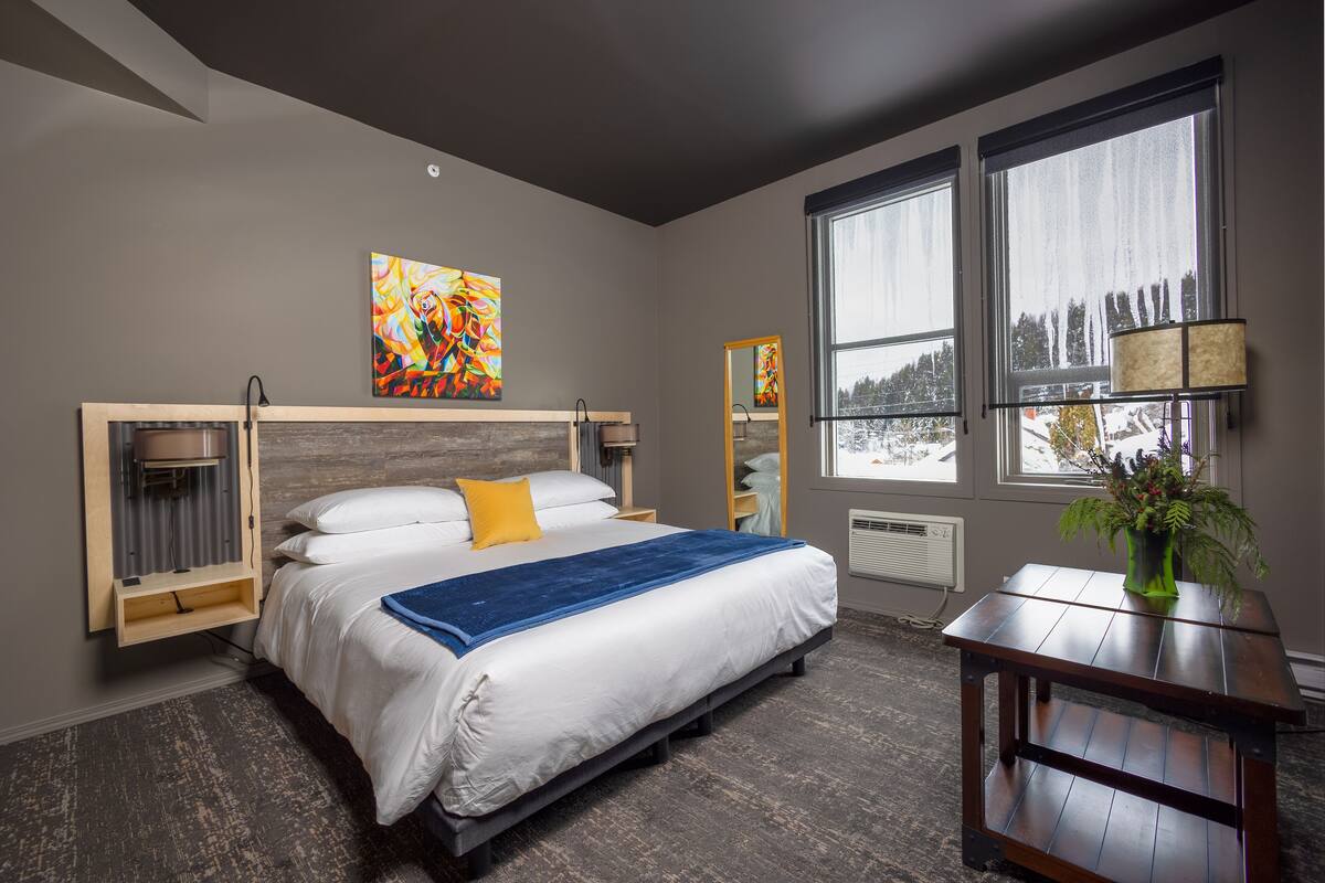 A modern bedroom with a bed, a wooden bedside table, a mirror, and a window with a view of trees.