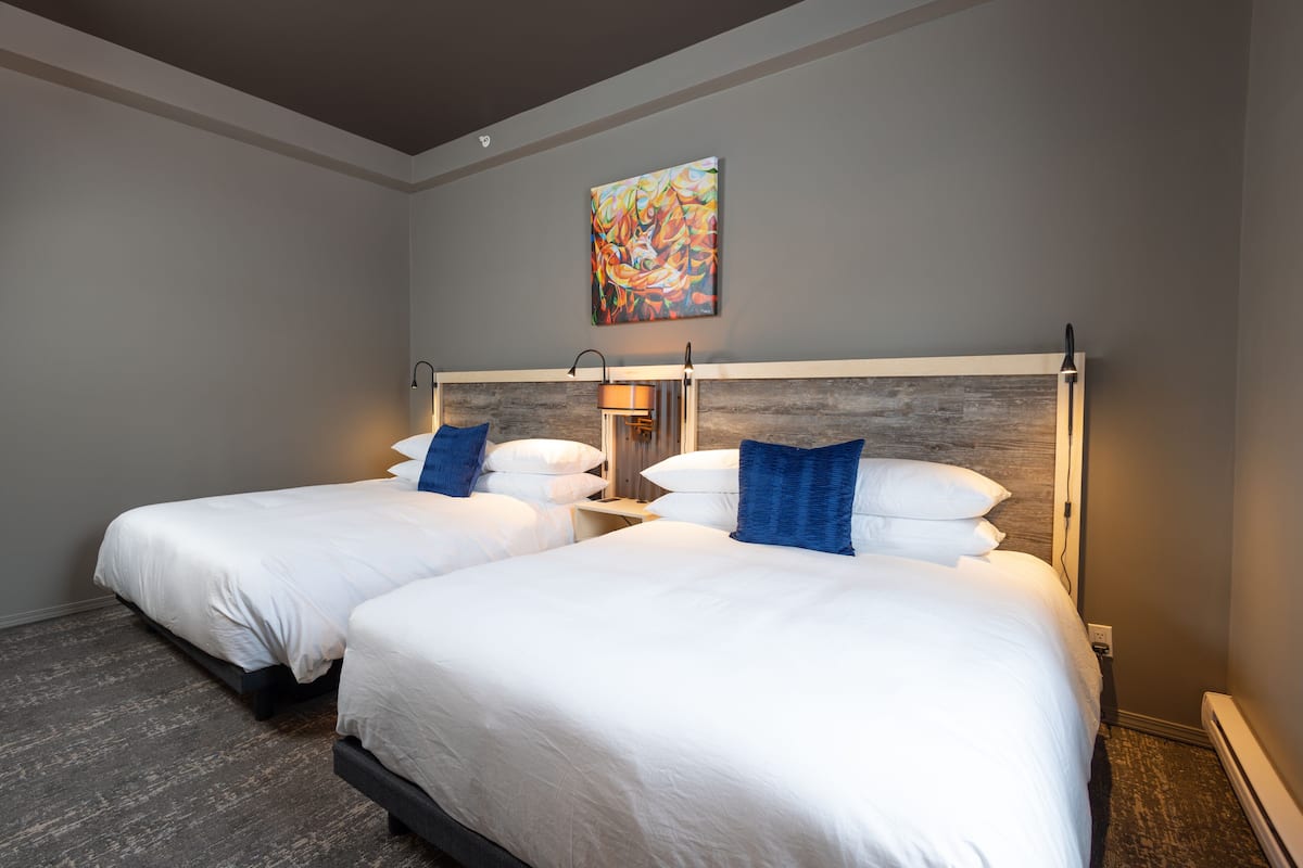 A hotel room with two beds, a wooden headboard, a painting on the wall, and a nightstand with a lamp.