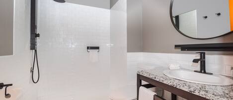 Un baño moderno con ducha envuelta en cristal, paredes de azulejos blancos, un lavabo blanco con grifo negro y un espejo redondo sobre el lavabo.