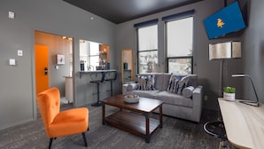 Una sala de estar moderna con un sofá gris, un sillón naranja y una mesa de centro de madera. Un televisor de pantalla plana está montado en la pared y una lámpara sobre una mesita auxiliar.