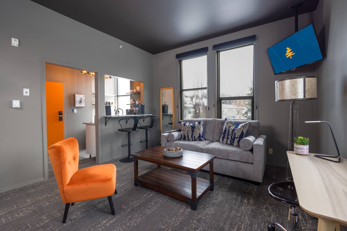 A modern living room with a grey sofa, orange armchair, and a wooden coffee table. A flat-screen TV is mounted on the wall, and a lamp is on a side table.