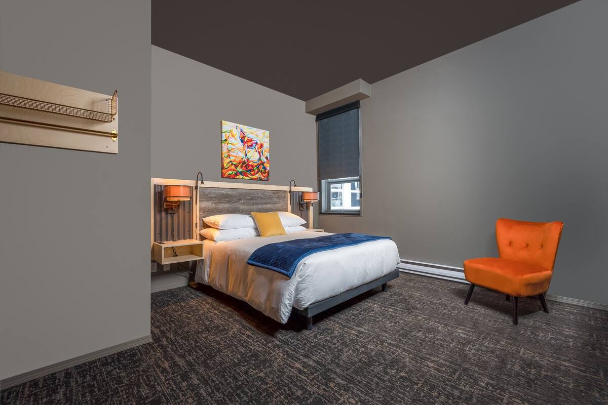 A modern hotel room with a large bed, a chair, a painting, and a window.