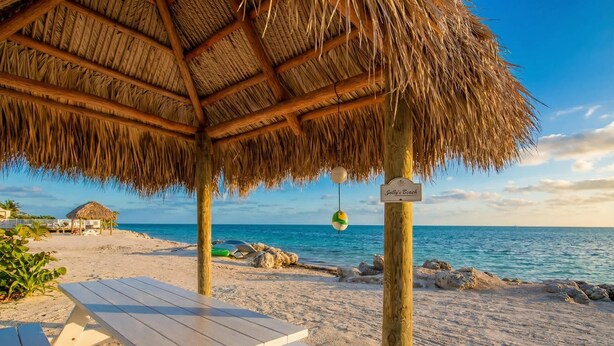 Una playa con una estructura de techo de paja, un letrero que dice 'Jolly's Beach' y una mesa de picnic.