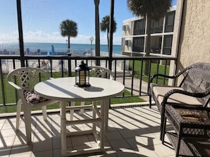 A balcony with a round table and two chairs, a lantern, and a view of the ocean and palm trees.
