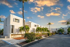 Un bâtiment moderne à la façade blanche et épurée, doté d’un parking et bordé de palmiers.