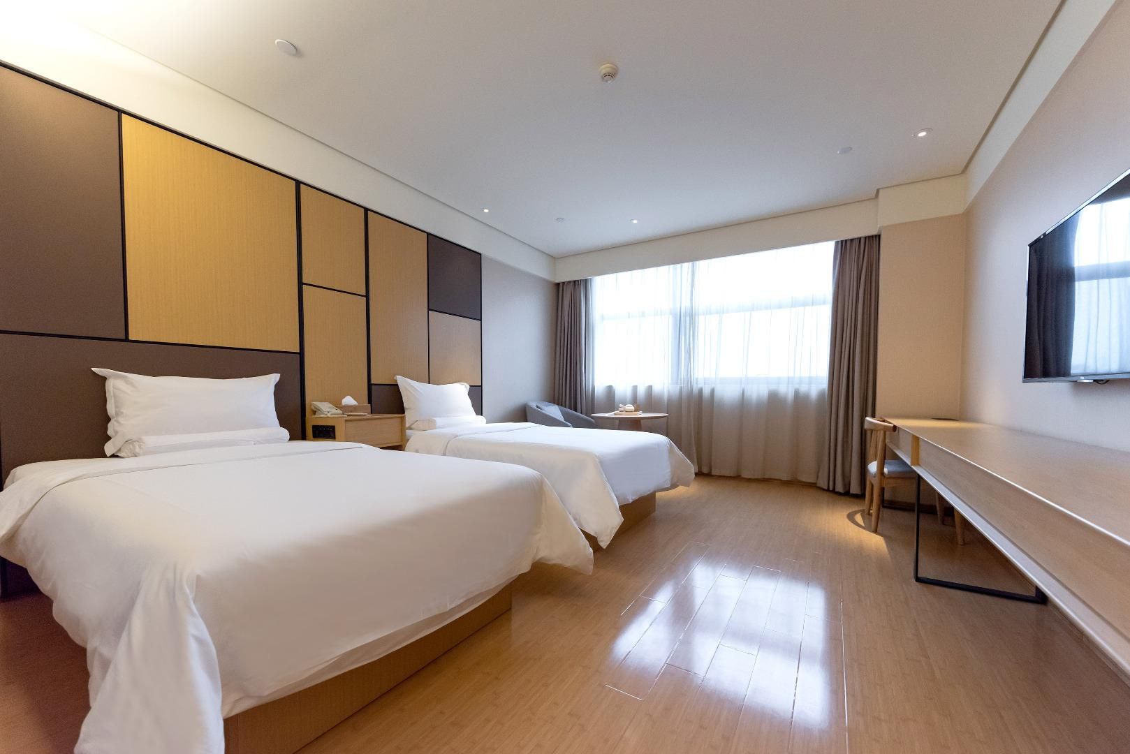 A hotel room with two beds, a desk, a TV, and a large window with curtains.