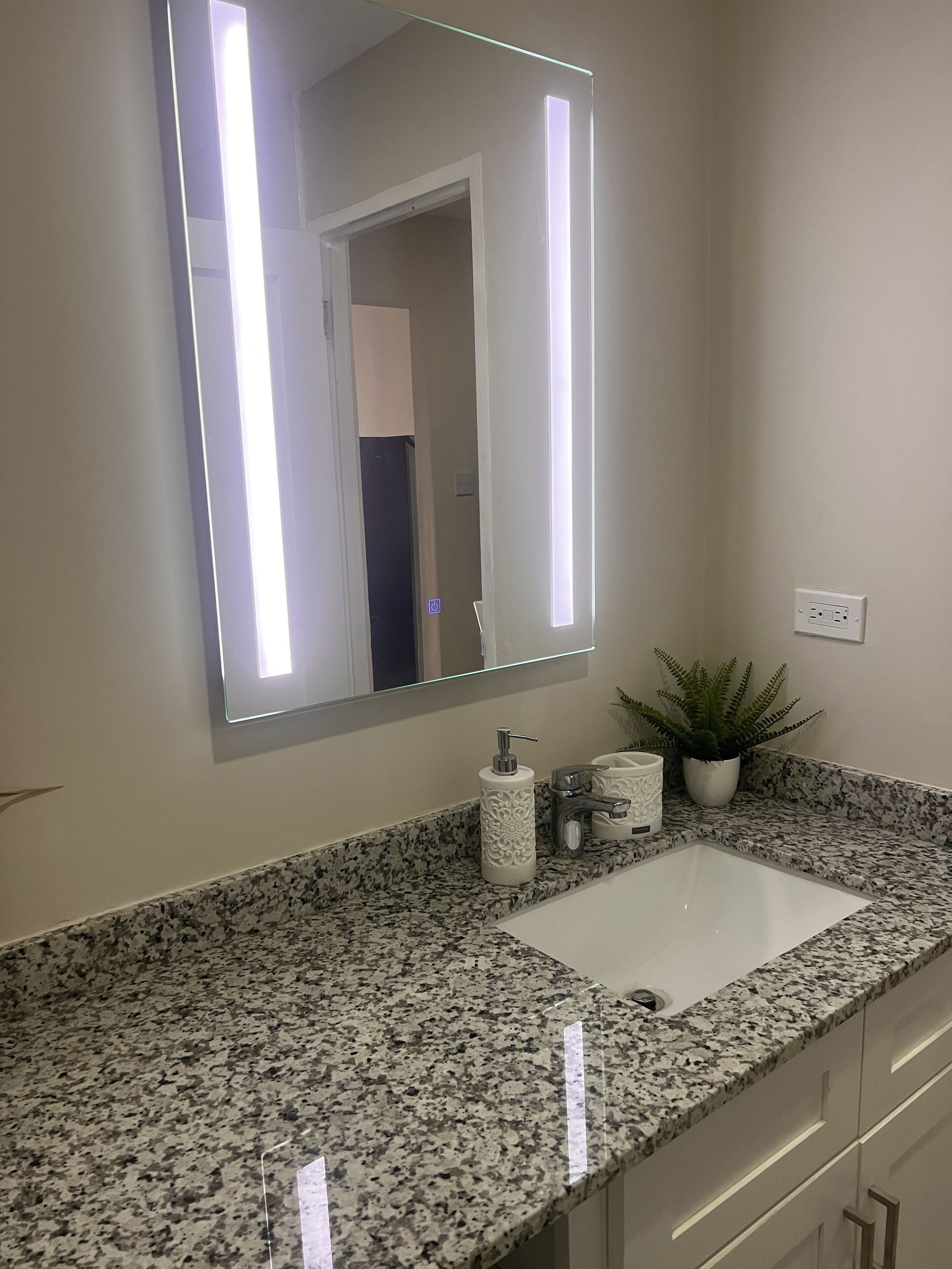 A bathroom with a granite countertop, a white sink, a large mirror with LED lights, a potted plant, and a towel dispenser.
