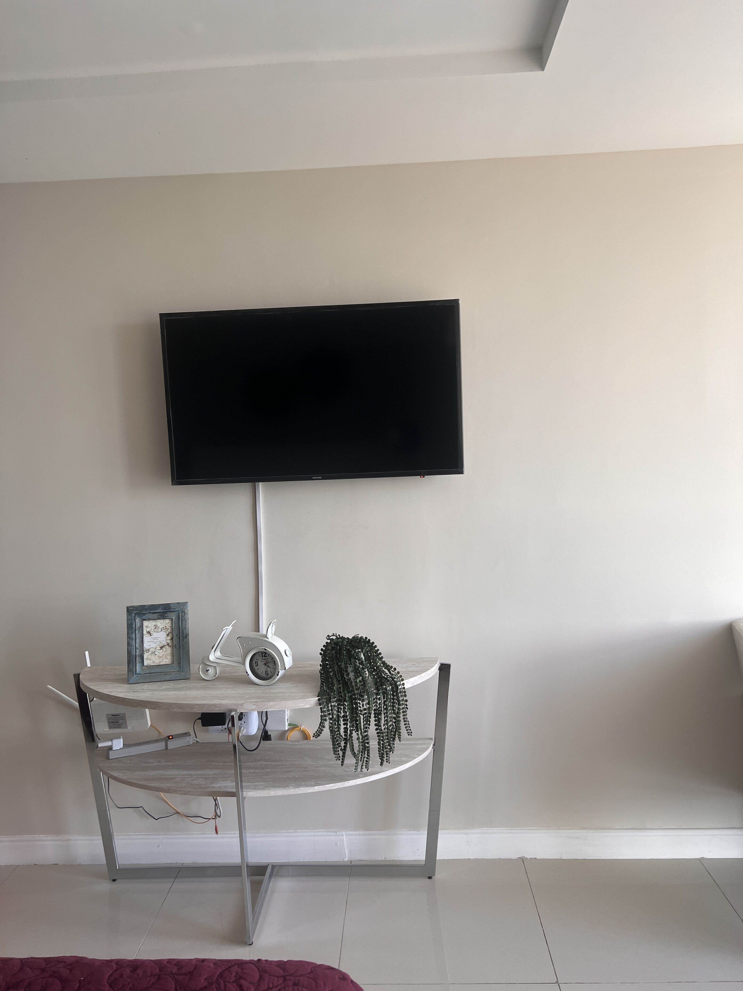A room with a flat-screen TV mounted on the wall, a small table with a clock and a framed picture, and a potted plant.