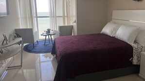 A hotel room with a large bed, a desk with a chair, a small table with two wine glasses and a plant, and a view of the sea through the window. - Sea Salt escape/ Beachfront apartment (Portmore)