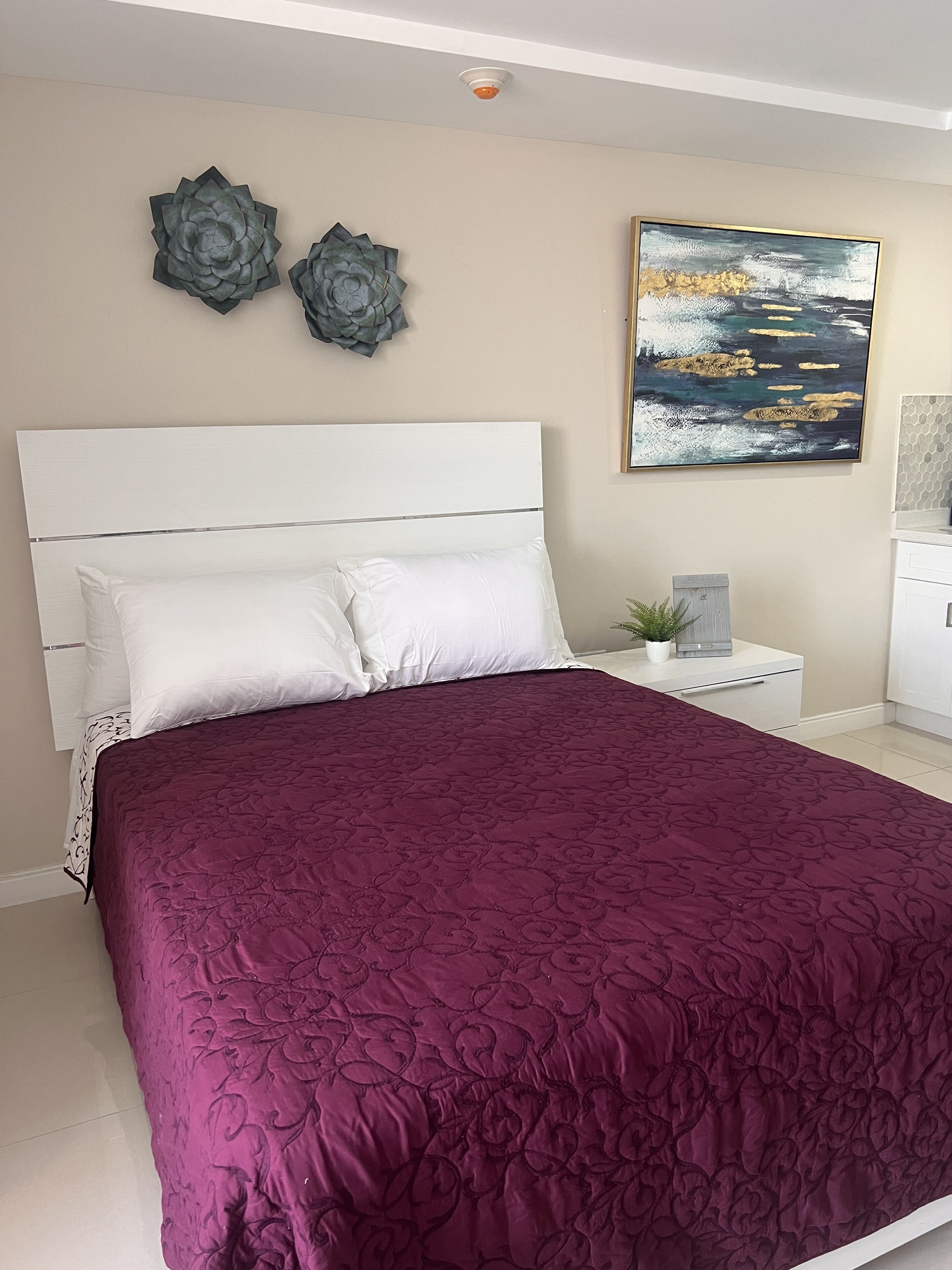 A bedroom with a large bed, white headboard, and a purple bedspread. There are two wall decorations and a painting above the bed.