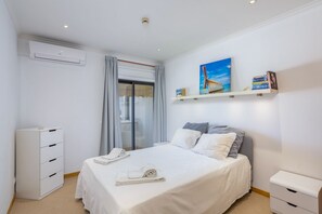 A bedroom with a bed, a bedside table, a wardrobe, a window with curtains, a painting, and a shelf with books. - Clube do Monaco Cozy Sea View 1-bed Apartment in Heart of Town (Albufeira)