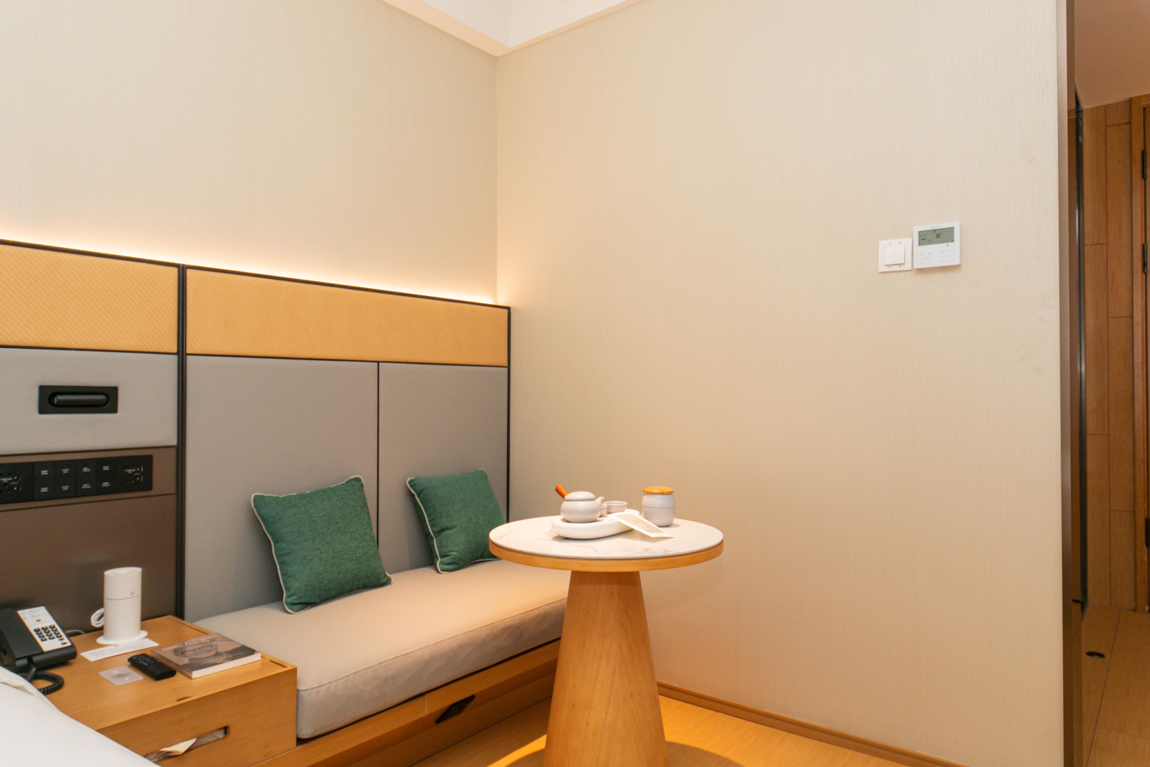 A hotel room with a bed, a bedside table with a phone and a book, a small round table with a teapot and cups, and a wall-mounted thermostat.