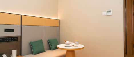 A hotel room with a bed, a bedside table with a phone and a book, a small round table with a teapot and cups, and a wall-mounted thermostat.