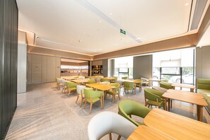 A modern dining area with multiple tables and chairs, a kitchen in the background, and large windows providing natural light. - Ji Hotel (Hangzhou)