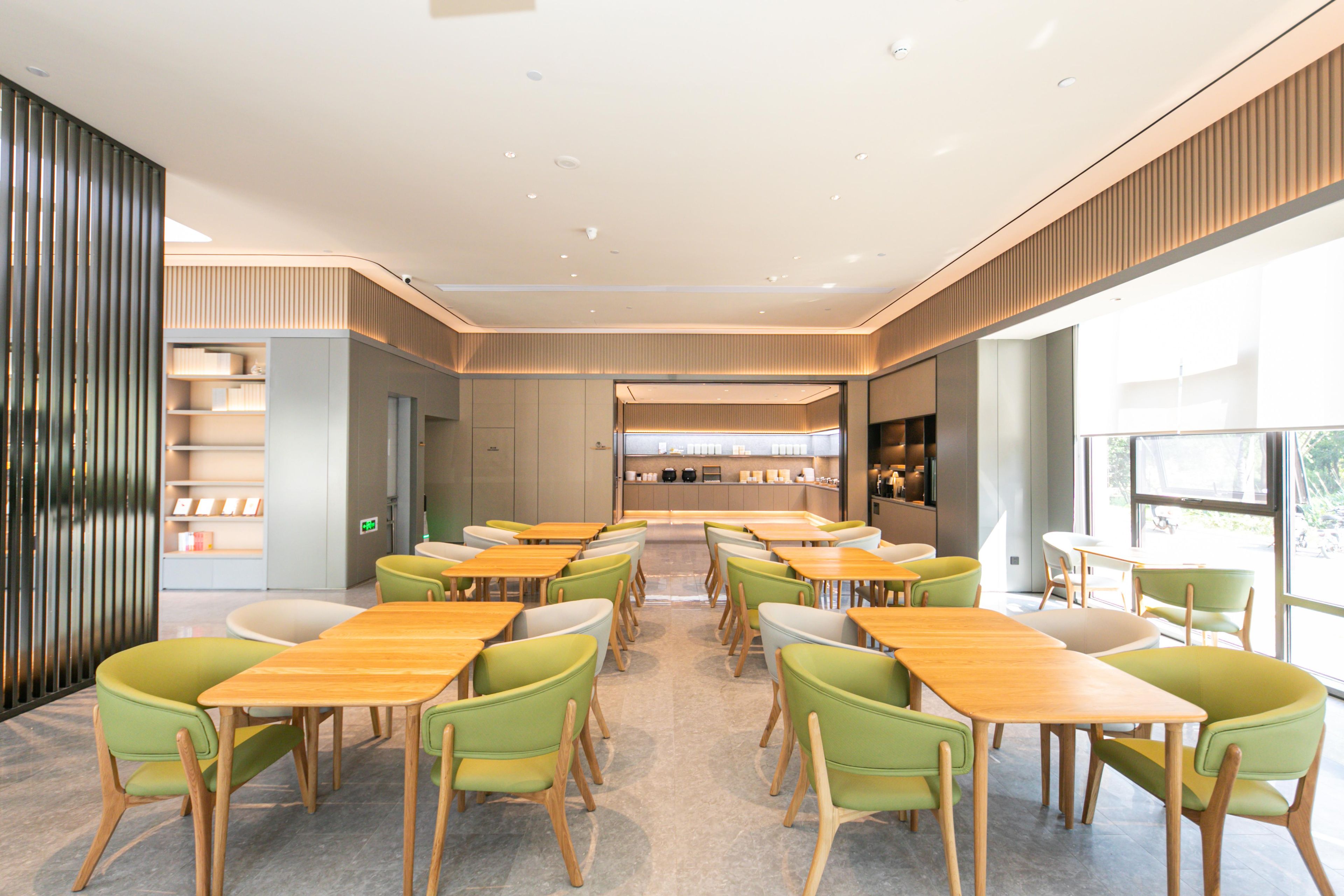 A modern dining area with wooden tables and green chairs, a kitchen in the background, and large windows allowing natural light.