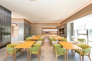 A modern dining area with wooden tables and green chairs, large windows, and a kitchen in the background. - Ji Hotel (Hangzhou)