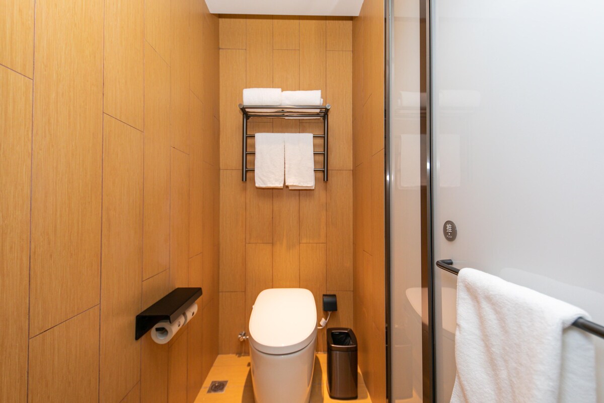 A compact bathroom with a toilet, a towel rack, a trash bin, and a glass shower enclosure.
