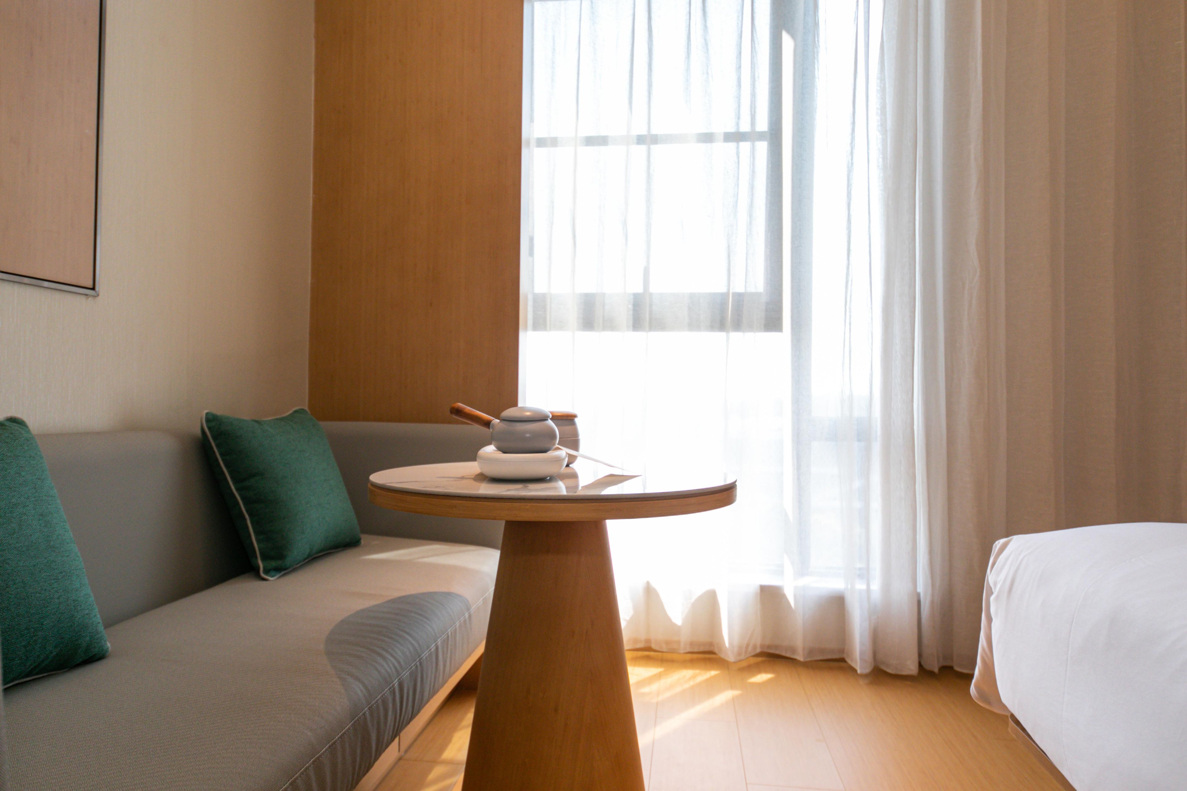 Een kamer met een houten tafel, een grijze bank, een wit bed en een raam met dunne gordijnen.