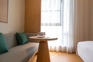 A room with a wooden table, a grey sofa, a white bed, and a window with sheer curtains. - Ji Hotel (Hangzhou)